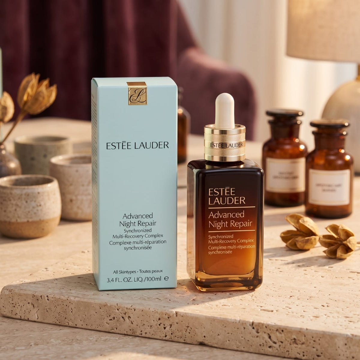 Estée Lauder Advanced Night Repair skincare set, featuring a light blue outer box and a dark amber glass dropper bottle with a gold collar, displayed on a stone tray with decorative botanical props.
