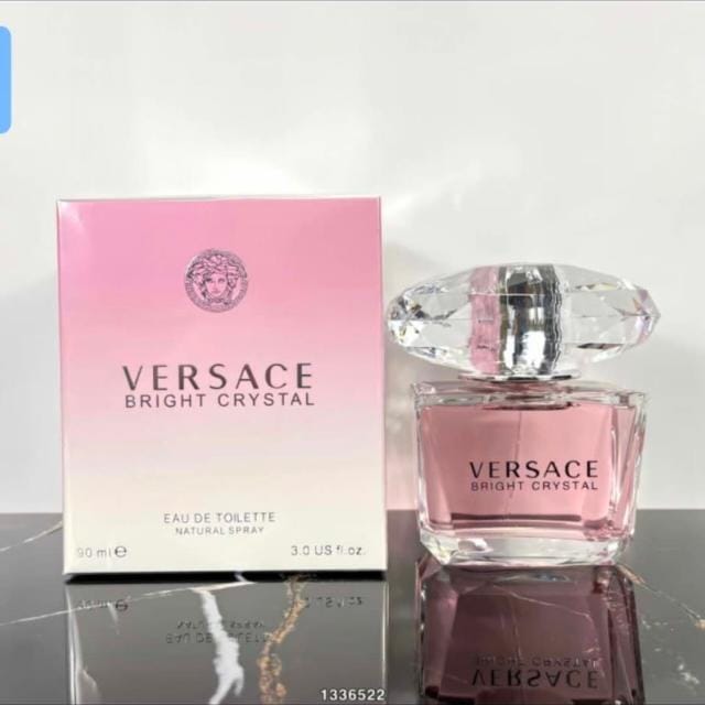 Versace Bright Crystal Eau de Toilette shown with its pink gradient box and the clear glass perfume bottle on a reflective dark surface.