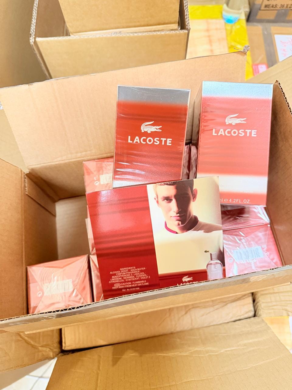 Several Lacoste perfume boxes, including a red and silver Eau de Toilette box and a pink box, stacked inside an open cardboard shipping box.